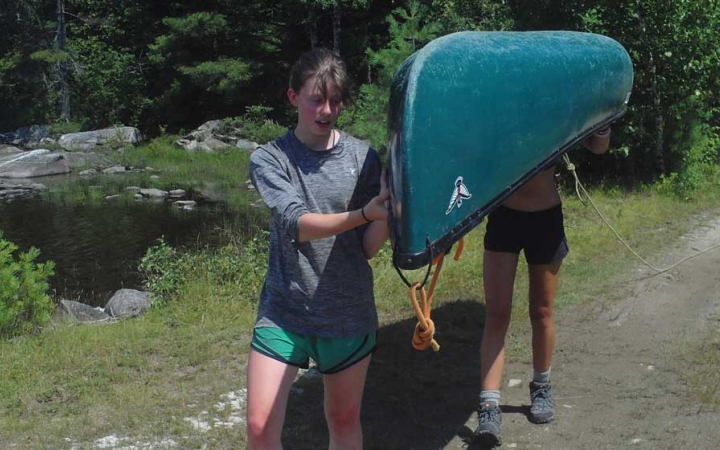 Two people carry an upside-down canoe on a trail in a wooded area